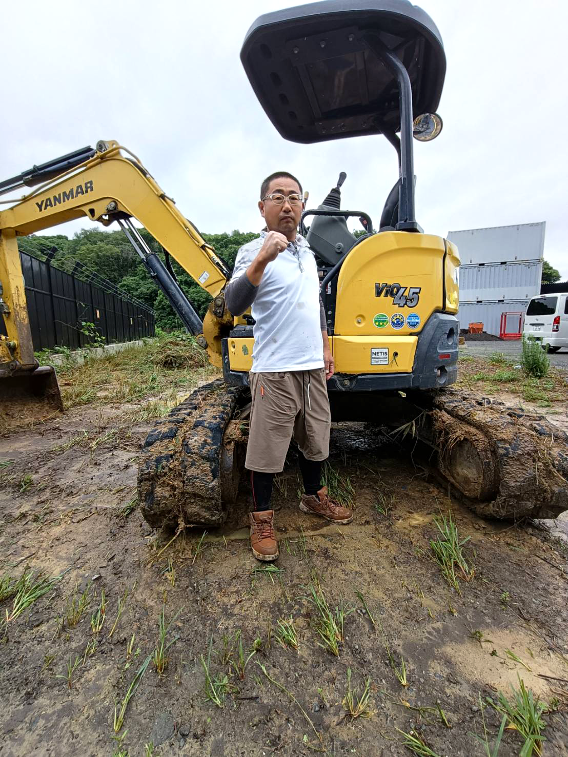 福津市のピース建設代表の出口昌央さんが、バックホウ（ショベルカー）を背景に腕組みをしている写真。